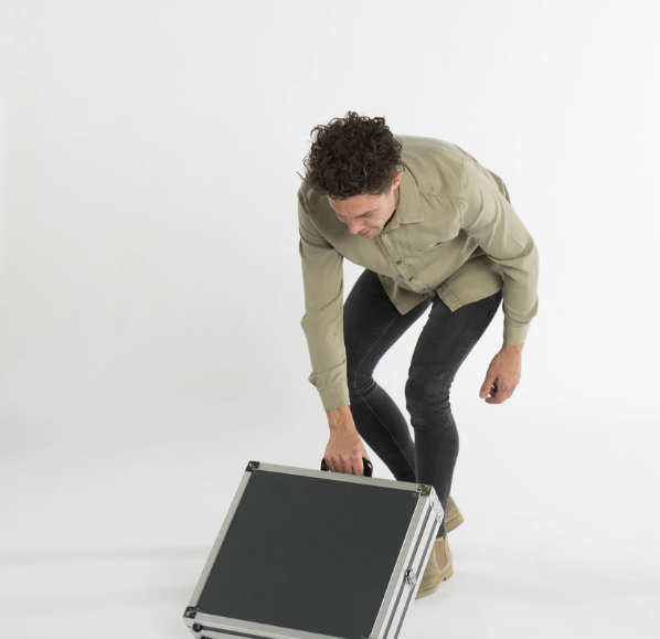 Man bukt zich om een opgevouwen, kofferachtige werktafel met handgreep op te tillen tegen een witte achtergrond.